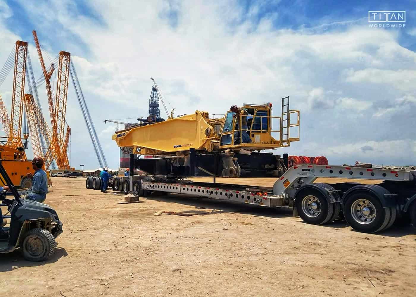 A disassembled Demag crane body loaded on a lowboy RGN trailer at an industrial job site, with crew members preparing the heavy equipment for transport near large crawler cranes and port operations.