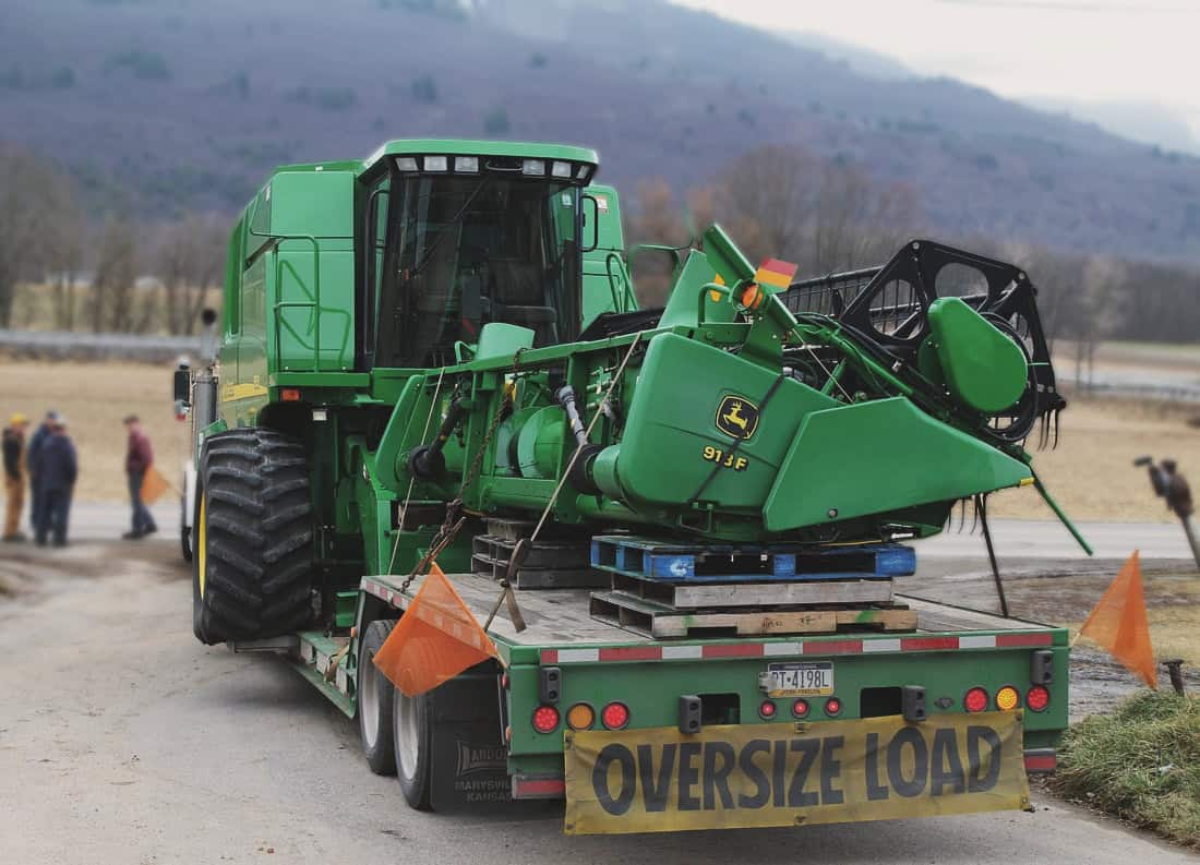 A John Deere 913F combine harvester loaded on a lowboy RGN trailer with “Oversize Load” signage, securely chained for transport through a rural farming area with rolling hills in the background.