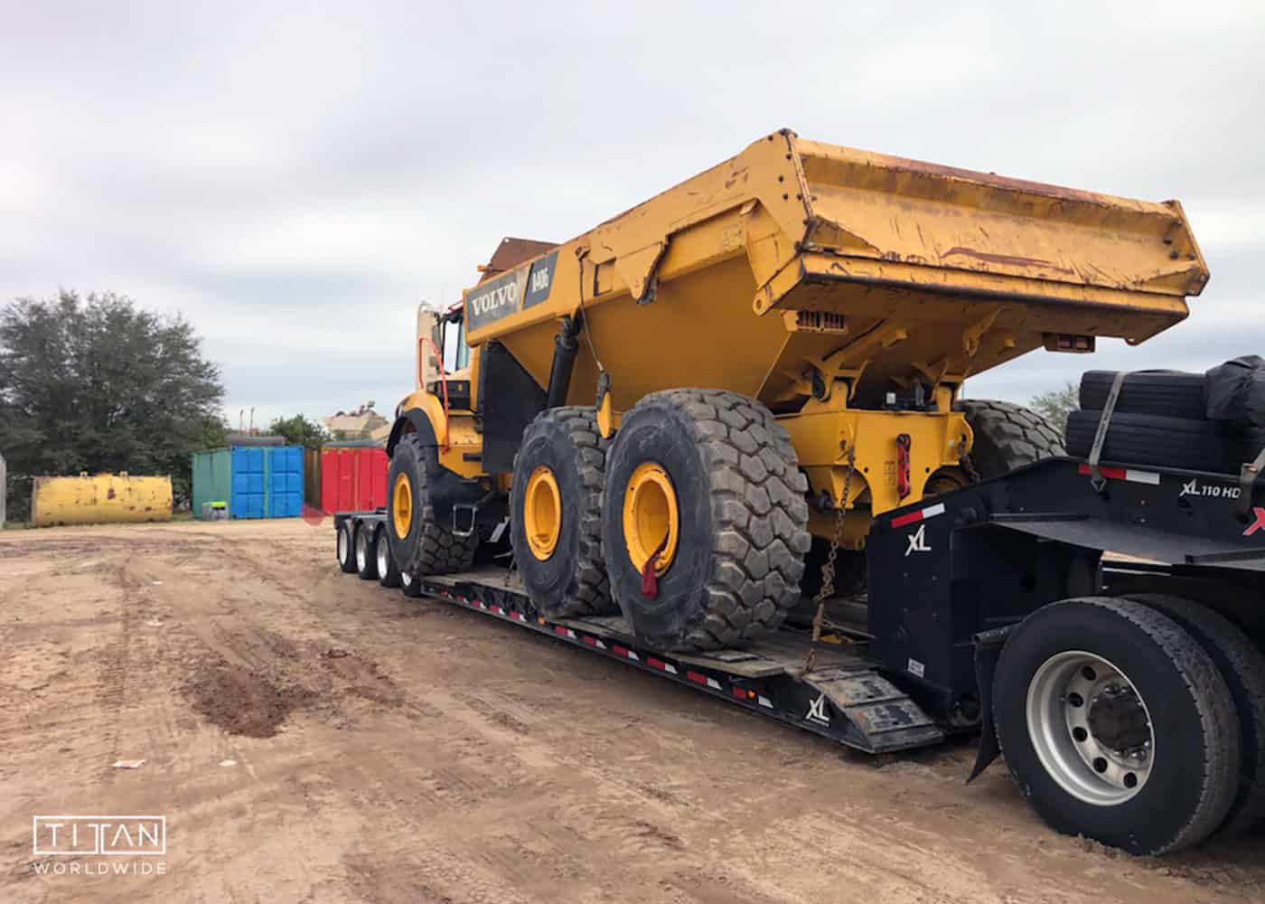 A Volvo A40G articulated dump truck loaded on an XL Specialized lowboy RGN trailer, secured for heavy equipment transport at a construction site with containers and machinery in the background.