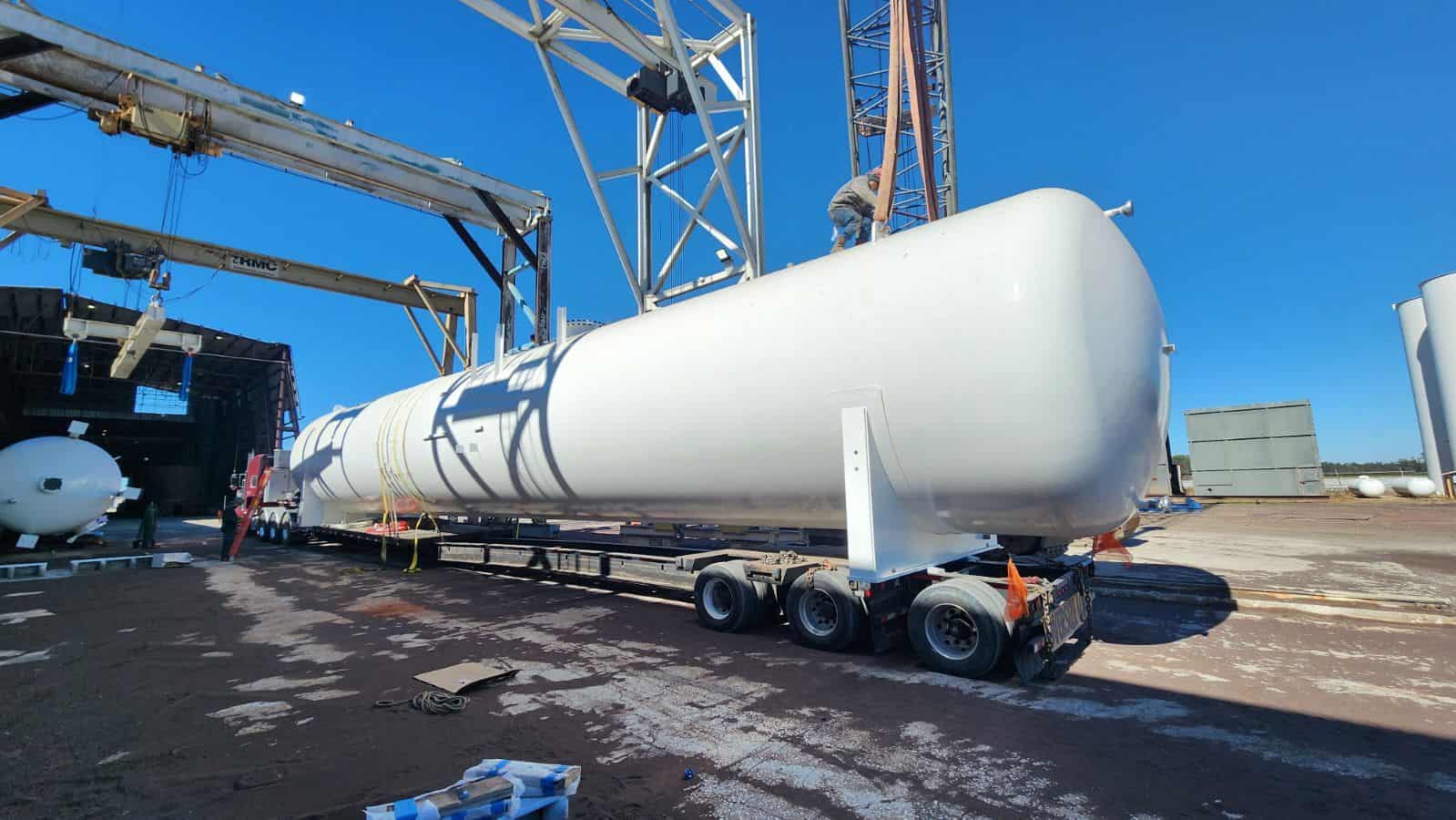 A large horizontal sulphuric acid tank being carefully loaded onto a multi-axle step-deck trailer using overhead cranes. Precision rigging and securement ensure safe handling of this oversized industrial vessel for over-the-road transport.