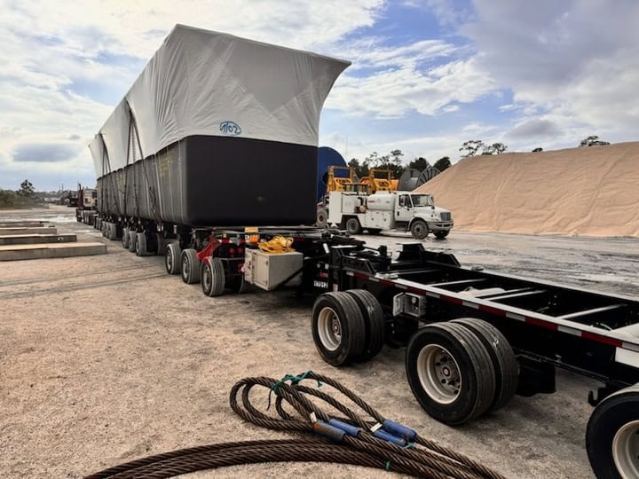 Specialized multi-axle heavy haul trailer hauling large industrial units covered with protective tarps at a construction or logistics yard, with support trucks and equipment visible in the background under partly cloudy skies.