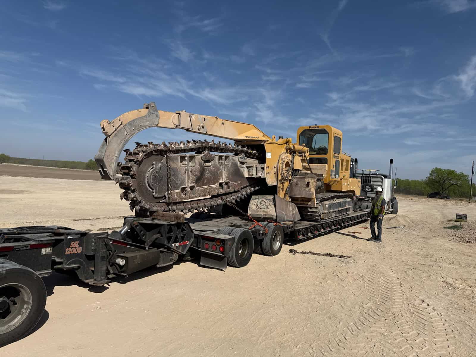 A Vermeer T858 trencher secured on an RGN lowboy trailer for transport across Texas. The photo highlights expert tie-down techniques, proper axle spacing, and specialized equipment handling for large pipeline and utility construction machinery.